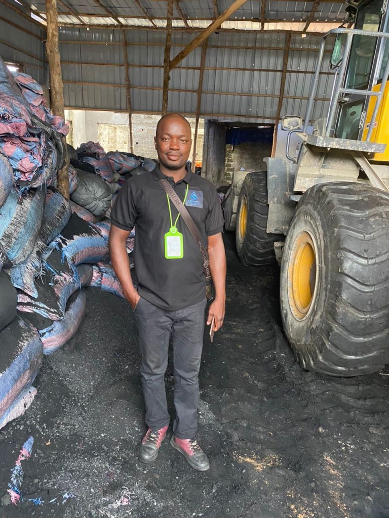 A man in warehouse full of bag of coals wearing black clothes with a tag tag on the neck beside a heavy duty truck.