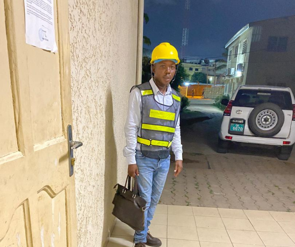 A man in a reflector working clothes with a hand bag and a helmet on in a yard.